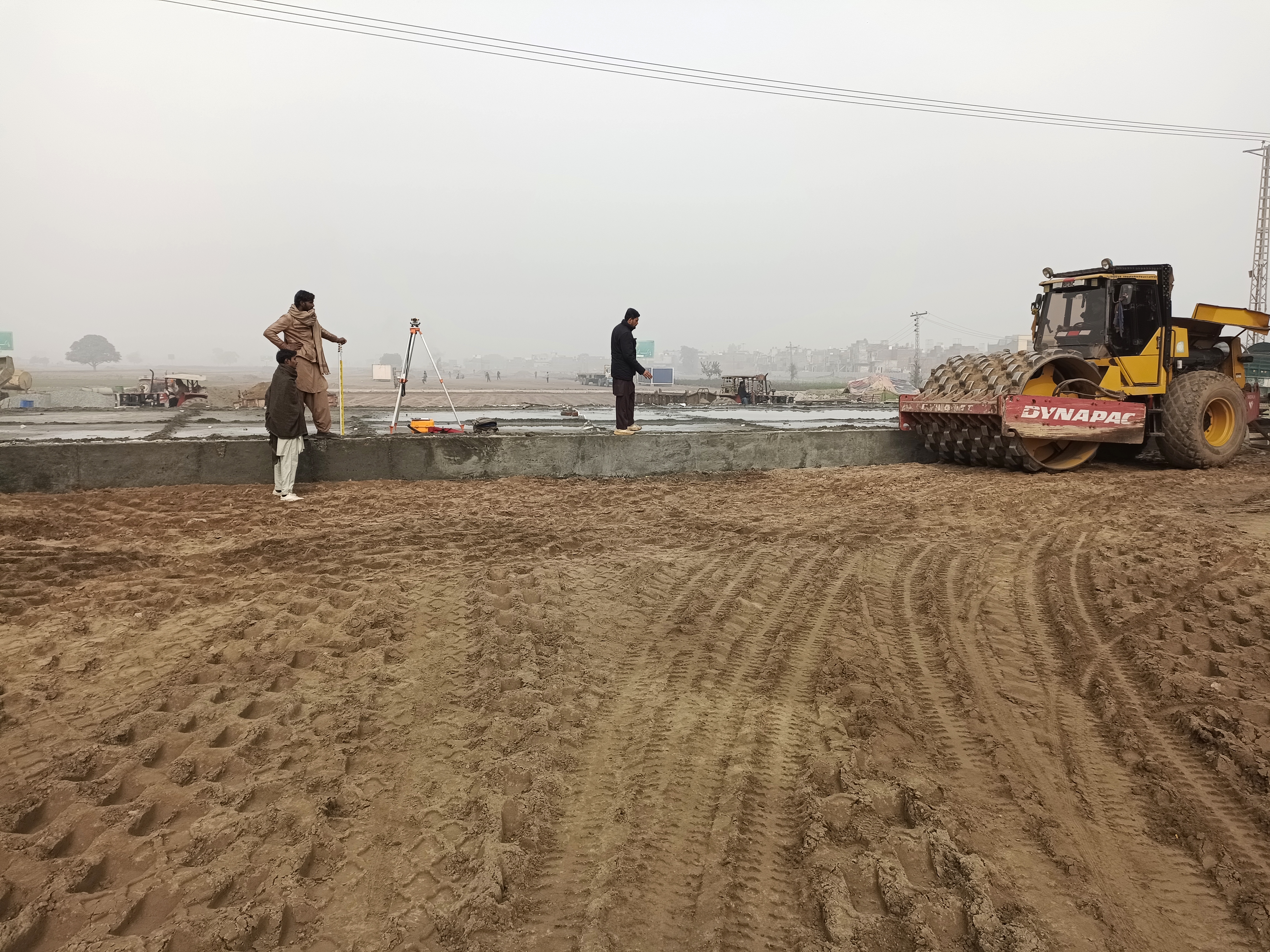 Two surveyors measuring with equipment, Dynapac roller in background, January 2023