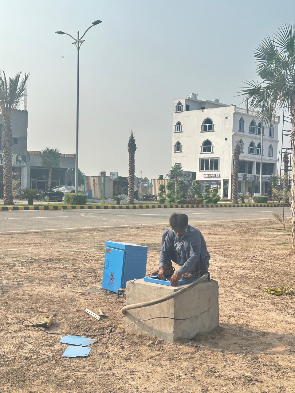 Worker servicing electrical junction box with palm-lined boulevard and white commercial building visible in background, September 2024